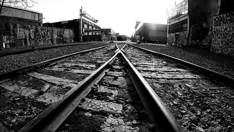 A black and white photo of two train tracks converging in an abandoned, graffeti-covered rail yard.